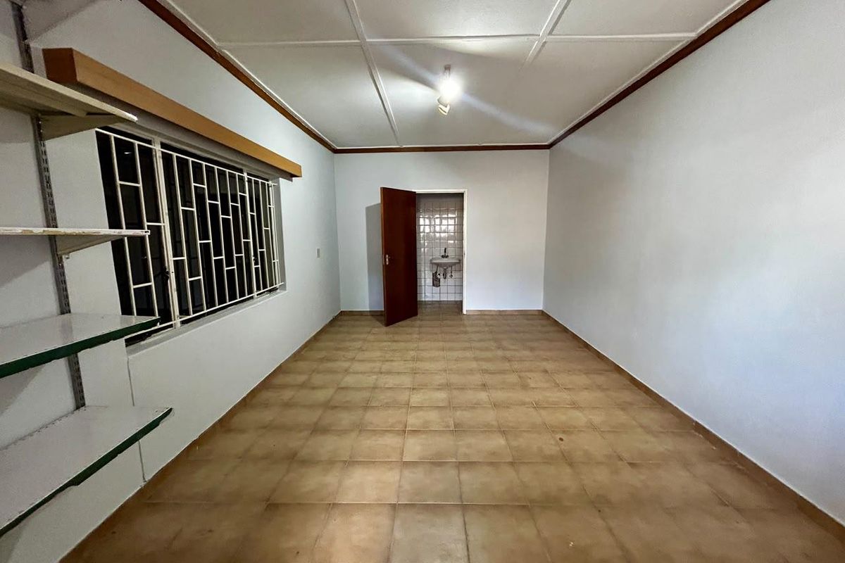 An empty, neutrally painted living area featuring tiled flooring, security bars on the window, and a view into an adjacent tiled utility space.
