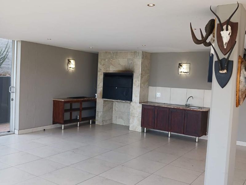 Spacious covered patio featuring a built-in braai with stone surround, fitted cabinetry with sink, tiled flooring, and direct access to the outdoors via sliding doors.