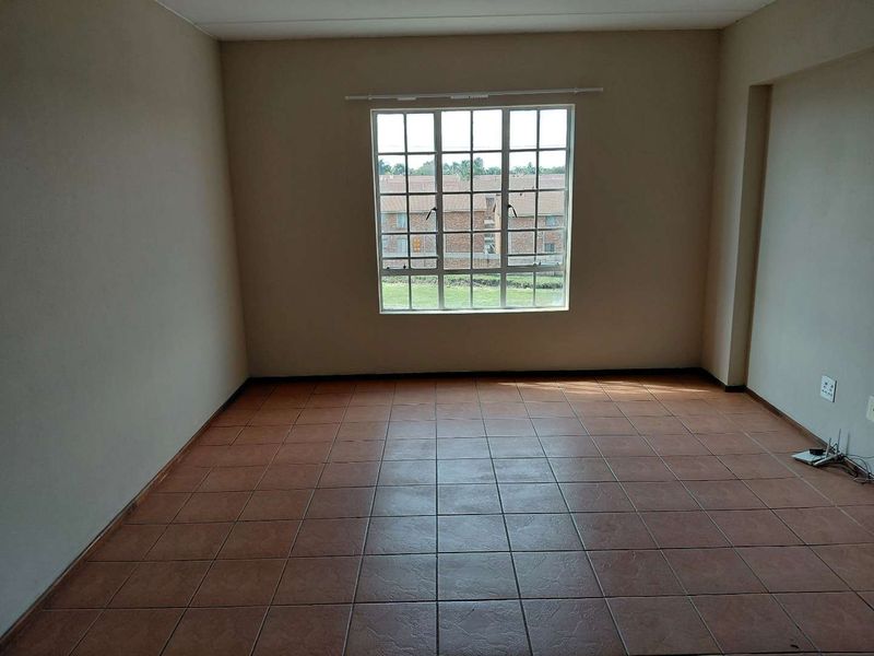 An empty living room featuring neutral walls, tiled flooring, and a large grid-pattern window providing natural light and a view of the complex grounds.