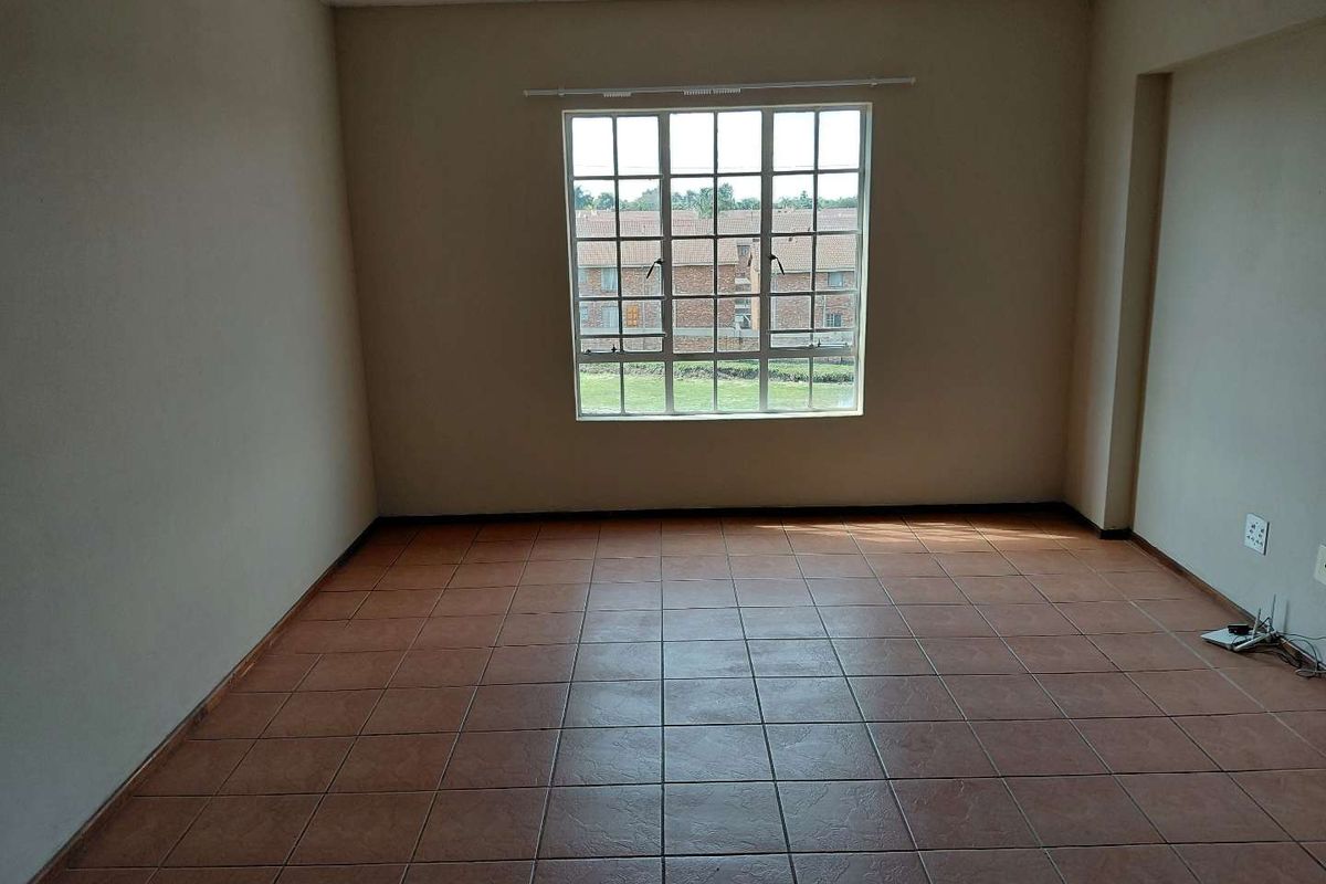 An empty living room featuring neutral walls, tiled flooring, and a large grid-pattern window providing natural light and a view of the complex grounds.