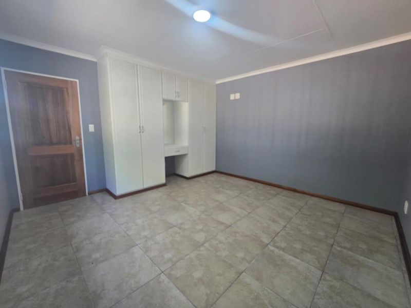 A spacious, empty bedroom featuring neutral tiled flooring, grey walls, and integrated white built-in cupboards with a central vanity area.