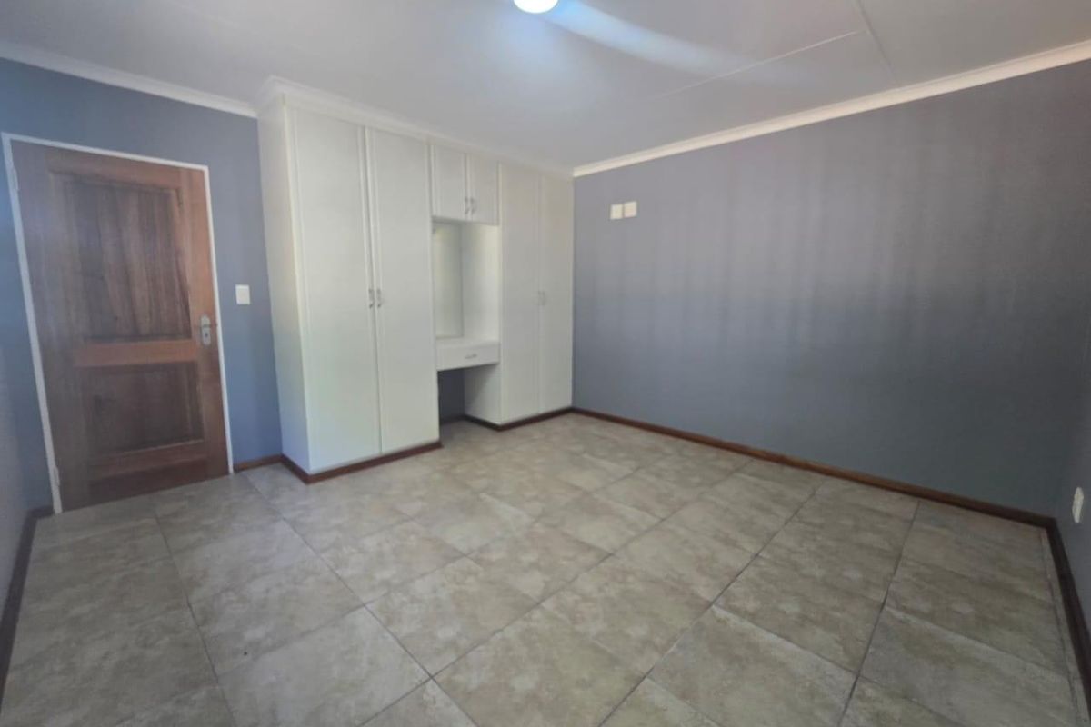A spacious, empty bedroom featuring neutral tiled flooring, grey walls, and integrated white built-in cupboards with a central vanity area.