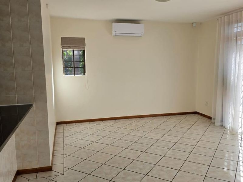 A bright, empty living area featuring neutral tiled flooring, an installed air conditioning unit, and large windows with fitted blinds.