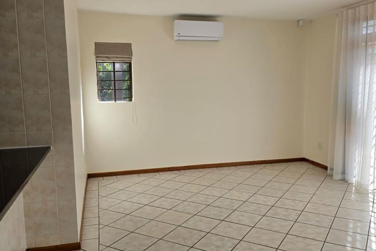 A bright, empty living area featuring neutral tiled flooring, an installed air conditioning unit, and large windows with fitted blinds.