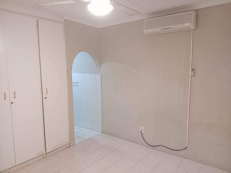 A clean, empty bedroom featuring tiled flooring, built-in cupboards, a ceiling fan, and a wall-mounted air conditioning unit.