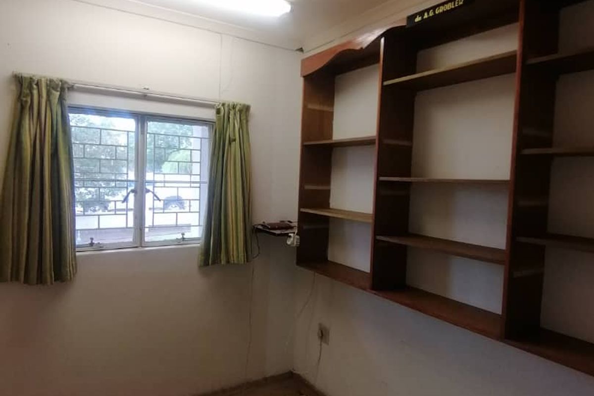 A small room featuring built-in wooden shelving, a window with security bars, and fluorescent lighting.