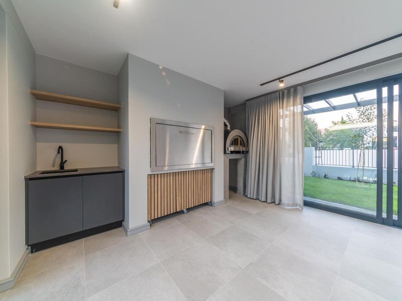 Modern covered patio featuring a built-in braai, pizza oven, kitchenette with sink, and large sliding doors opening to the garden.