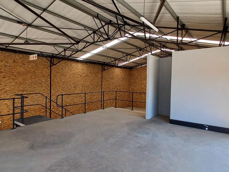 Spacious industrial mezzanine area featuring exposed brick walls, steel roof trusses, concrete flooring, and natural light from skylights.