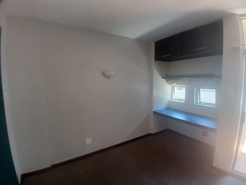 An empty bedroom featuring a built-in desk, overhead storage cabinetry, and wall-mounted lighting, providing a functional space for a home office or study area.
