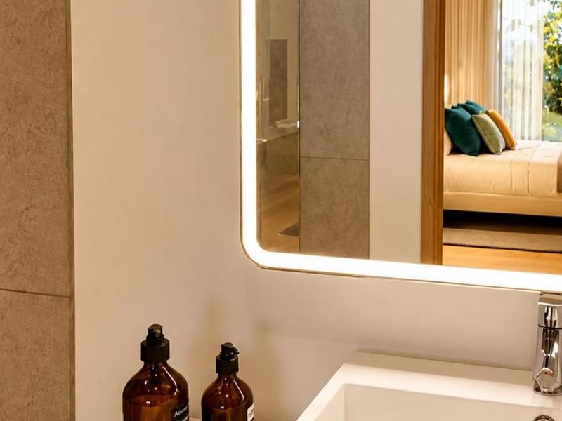 A modern bathroom vanity featuring a sleek basin, chrome faucet, and a contemporary backlit mirror, with a view into the adjacent bedroom.