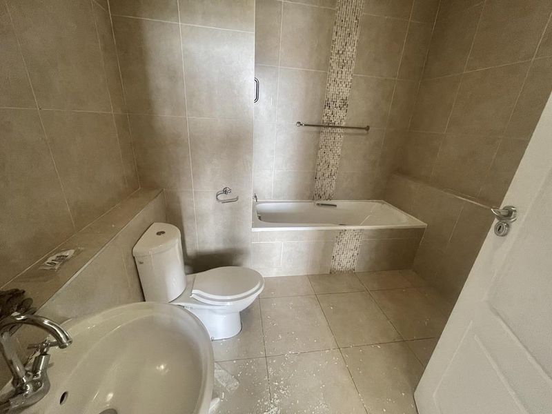 A neutral-toned bathroom featuring a built-in bathtub, pedestal washbasin, and dual-flush toilet, finished with floor-to-ceiling ceramic wall tiles.
