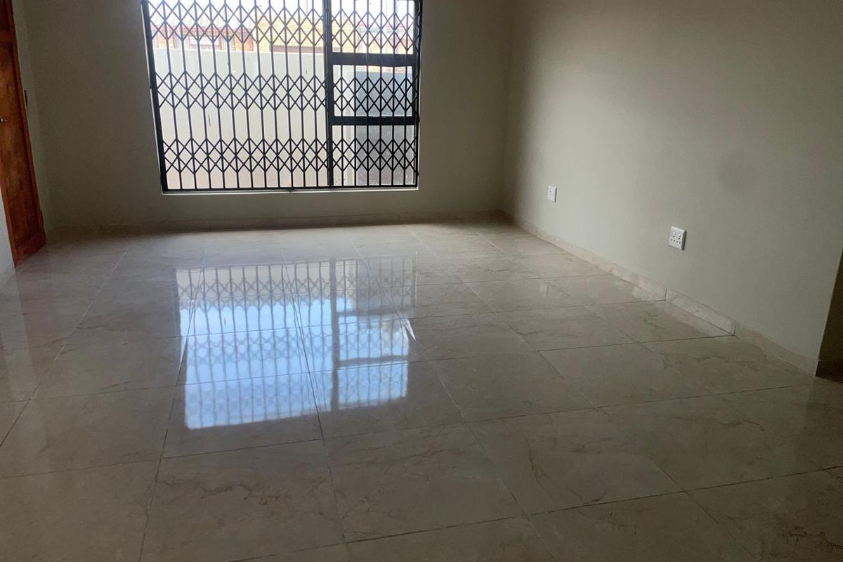 A bright, empty living room featuring polished porcelain floor tiles and a large window secured with a metal security gate.