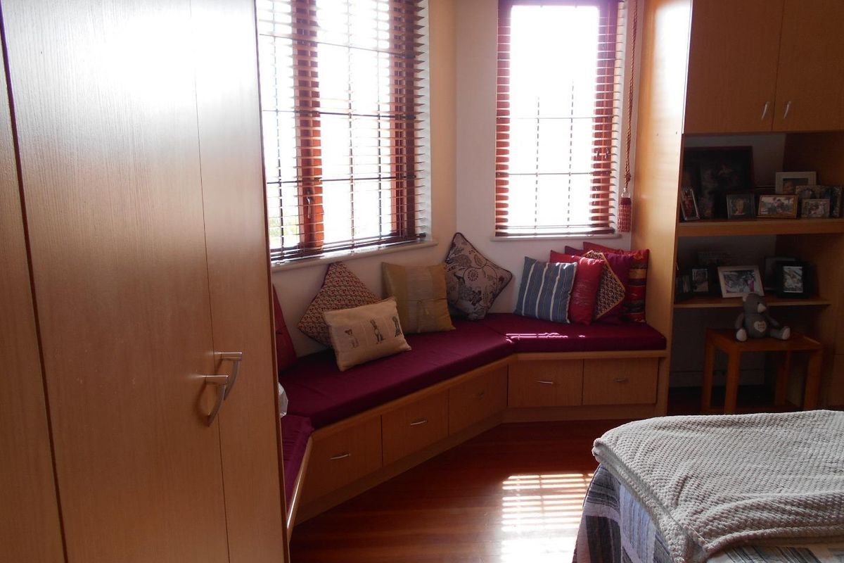A cozy bedroom featuring custom built-in wooden cabinetry, a corner window seat with integrated storage, and warm timber flooring.