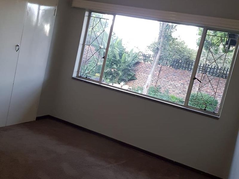 A vacant bedroom featuring built-in wardrobes, carpeted flooring, and large windows with security bars overlooking a rear garden area.