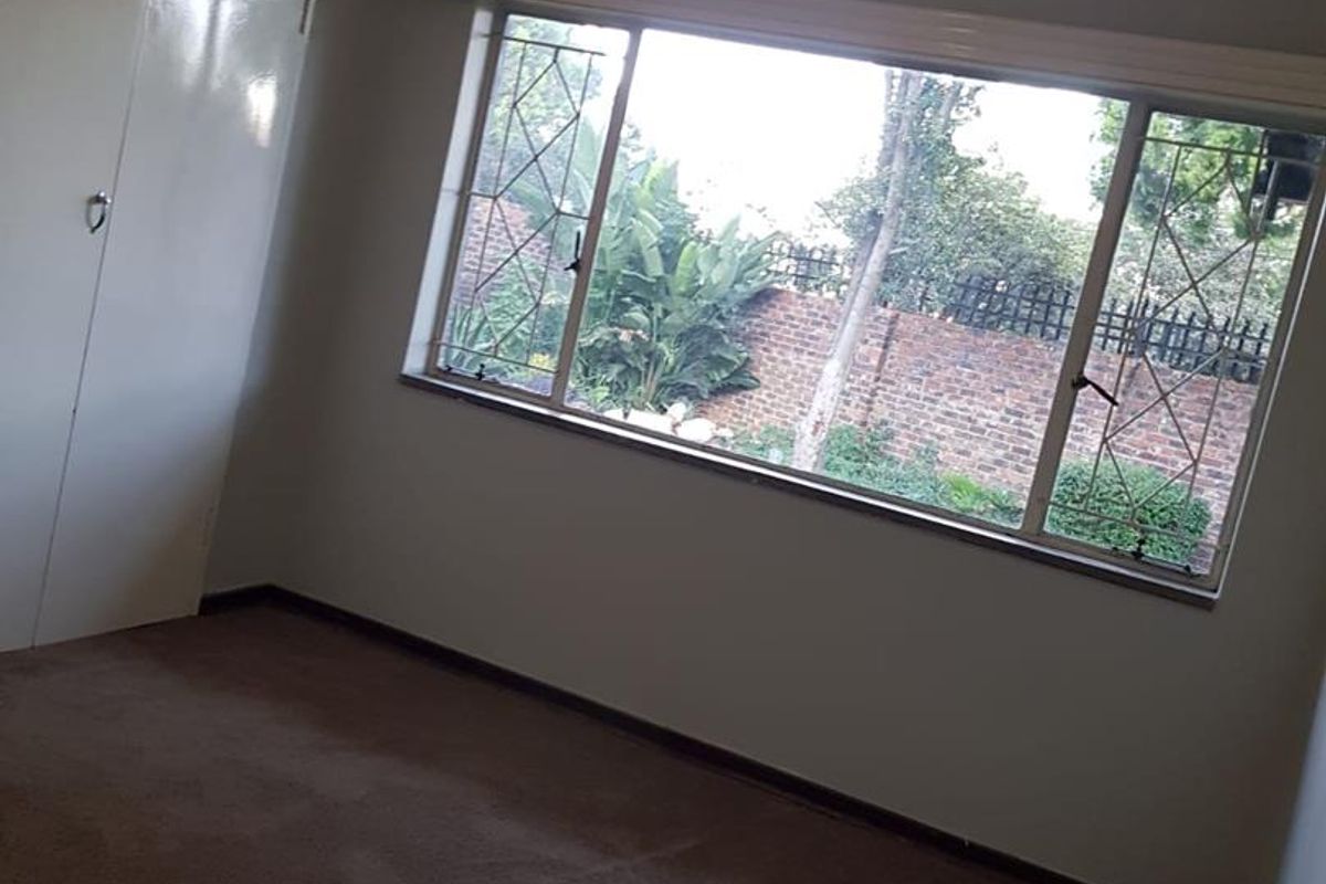 A vacant bedroom featuring built-in wardrobes, carpeted flooring, and large windows with security bars overlooking a rear garden area.