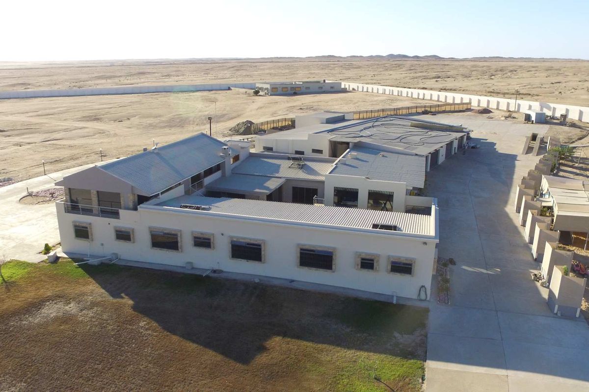 Aerial view of a large property complex featuring multiple connected buildings, extensive paved areas, and a perimeter wall in a desert landscape setting.
