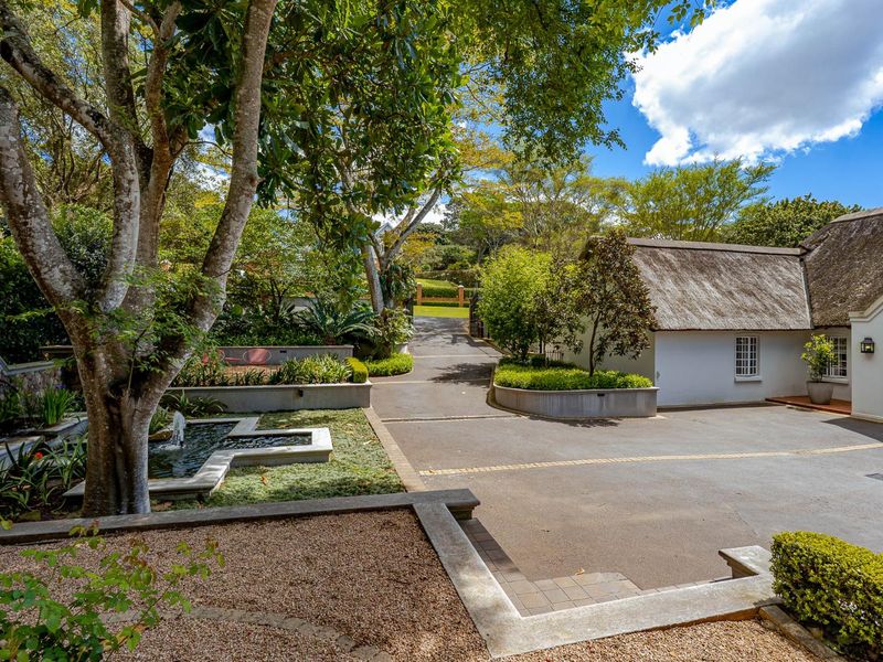 A beautifully landscaped front entrance featuring a paved driveway, a charming thatched-roof structure, and a tranquil water feature set within a lush garden.