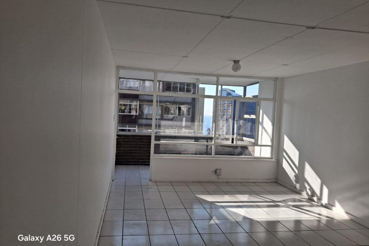 An empty, sunlit living area featuring tiled flooring and a large glass window wall with views of the surrounding apartment buildings.