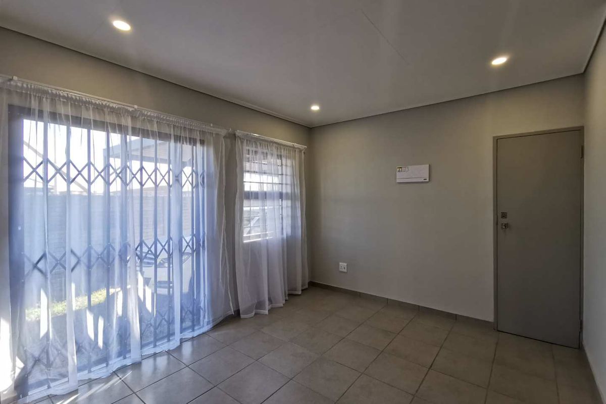 A bright, empty living room featuring neutral tiled flooring, recessed lighting, and a sliding glass door with security gate leading to the exterior.