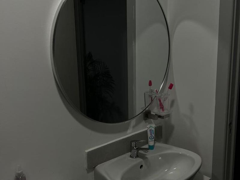 A functional bathroom featuring a white vanity cabinet with a ceramic basin, chrome faucet, and a circular wall-mounted mirror.