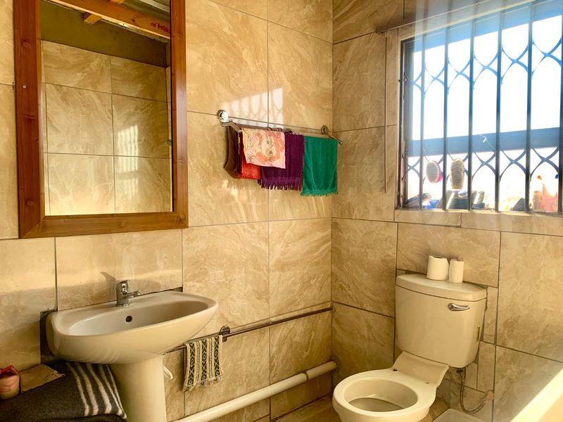 A functional bathroom featuring full-height wall tiling, a pedestal washbasin, a standard toilet, and a window fitted with security bars.