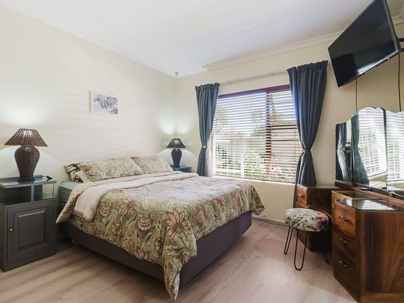 A well-lit bedroom featuring light-toned laminate flooring, neutral walls, and a window fitted with venetian blinds.