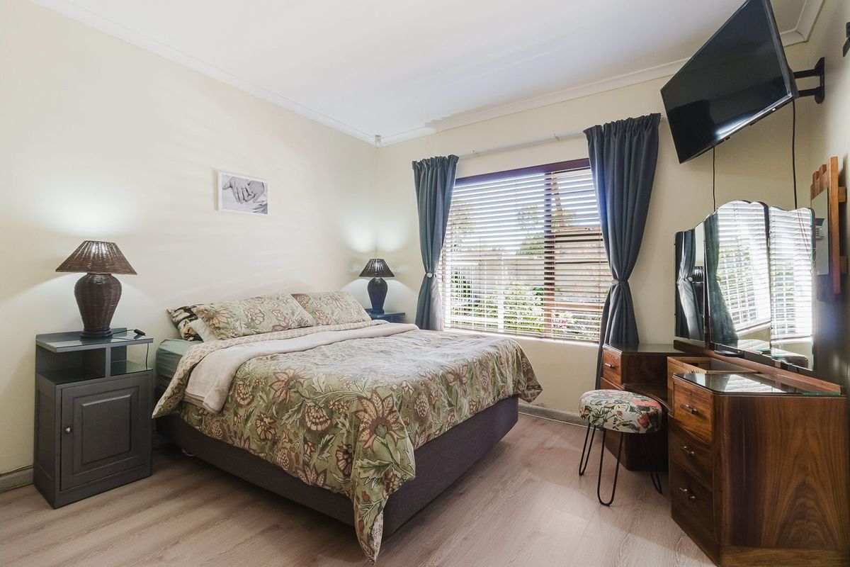 A well-lit bedroom featuring light-toned laminate flooring, neutral walls, and a window fitted with venetian blinds.