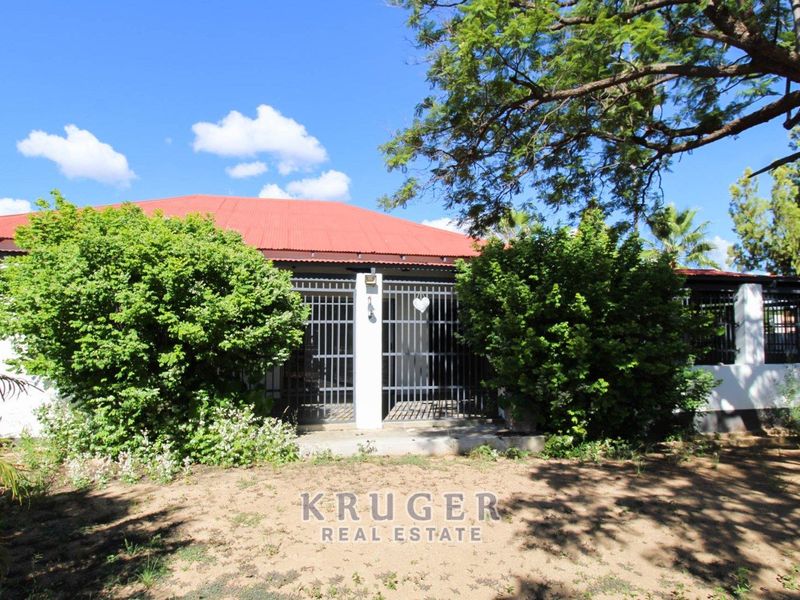 Front elevation of a house featuring a red corrugated metal roof, security gate entrance, and surrounding garden area.