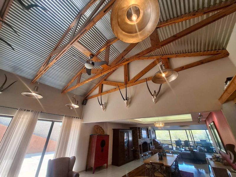 Spacious open-plan living area featuring high vaulted ceilings with exposed timber trusses, corrugated metal roofing, and a prominent brick fireplace.