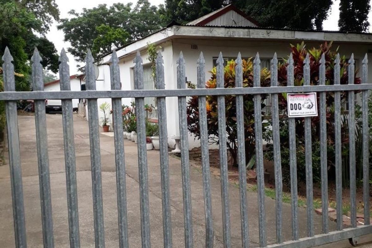 Front view of the property featuring a galvanized steel security gate, concrete driveway, and a single-story house with a corrugated metal roof.