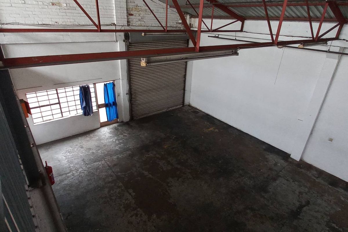 Spacious industrial warehouse unit featuring high ceilings with exposed steel trusses, a roller shutter door, and a mezzanine level overlooking the main floor area.