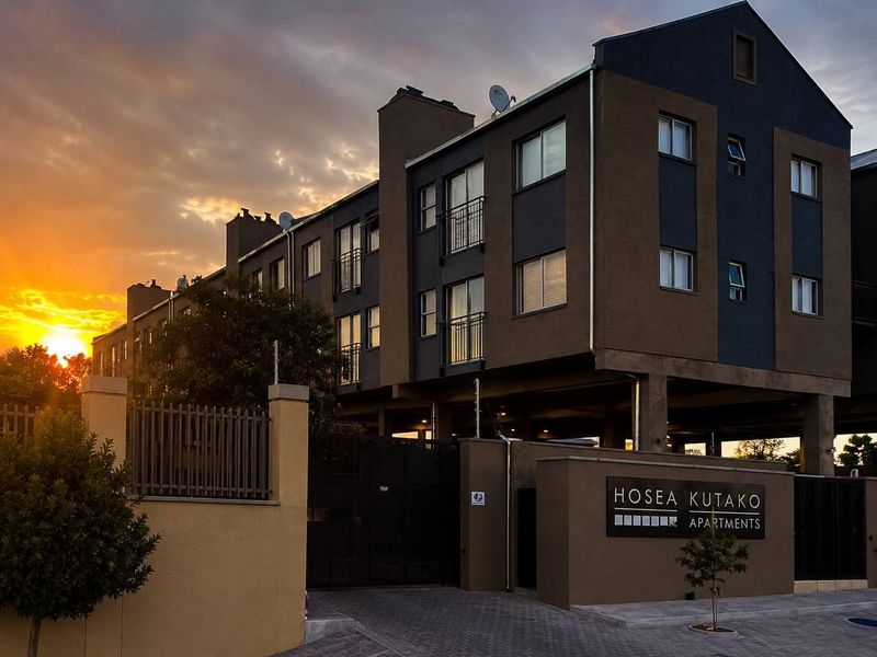 Exterior view of the Hosea Kutako apartment complex featuring modern facade, secure gated entrance, paved driveway, and balconies at sunset.