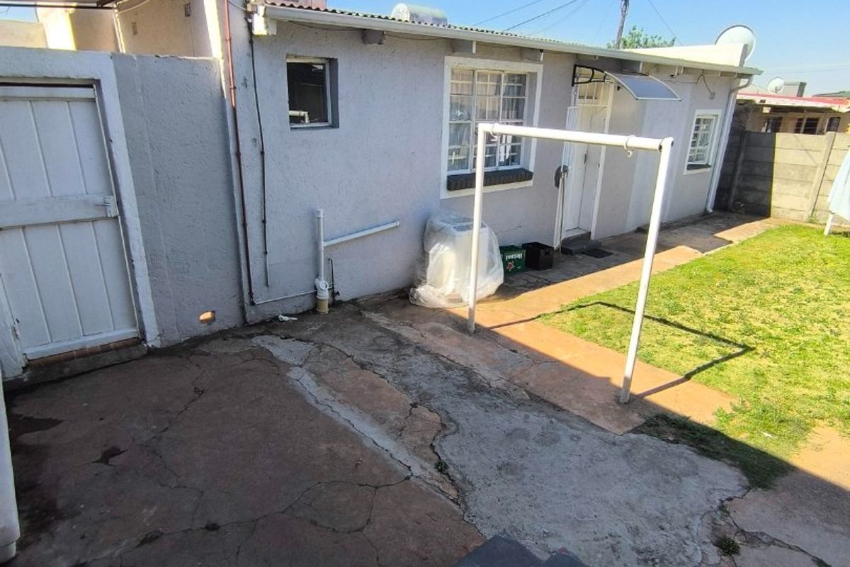 Rear exterior view featuring a concrete patio area, lawn, solar water heater, and boundary wall, showing signs of wear and aging.