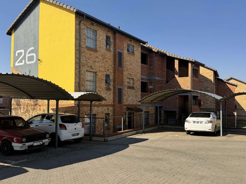 Exterior view of a multi-story face brick apartment building featuring covered parking bays and a paved driveway area.