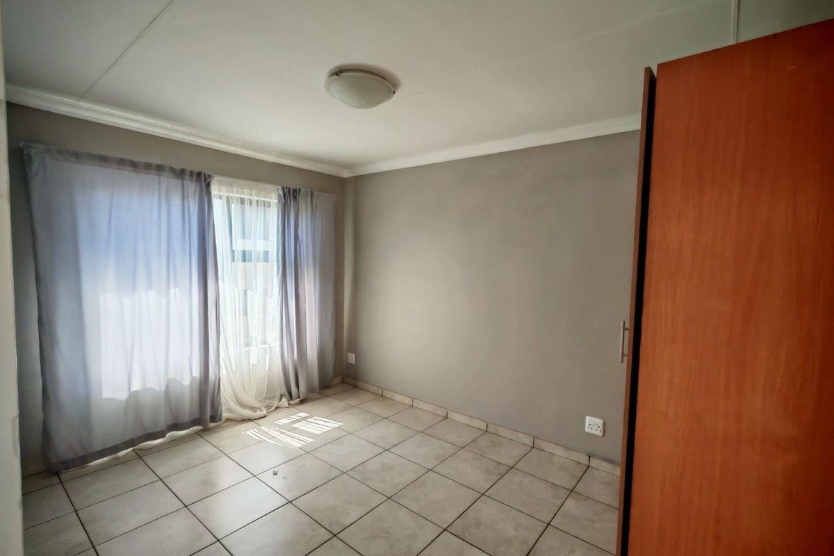 A bedroom featuring neutral grey walls, tiled flooring, a built-in wooden wardrobe, and large windows with curtains allowing natural light.