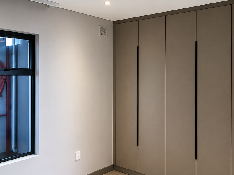 A modern, empty bedroom featuring neutral tiled flooring, recessed lighting, and sleek floor-to-ceiling built-in cupboards with contemporary black handles.