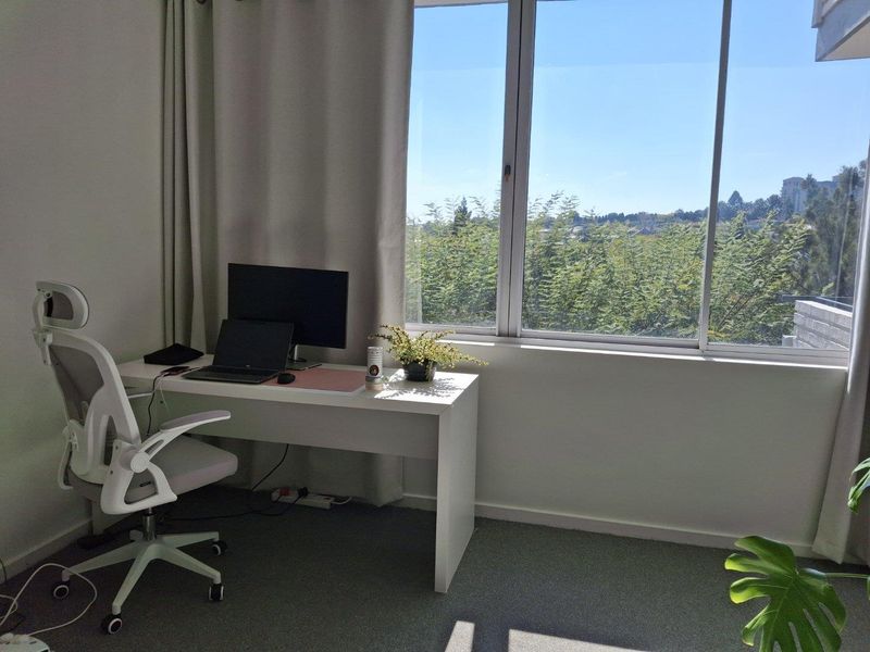 A bright home office space featuring large windows with natural light and carpeted flooring, suitable for a productive work environment.