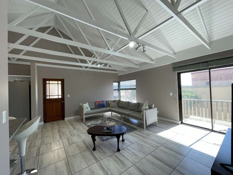 Spacious open-plan living area featuring a high vaulted ceiling with exposed trusses, ceramic tiled flooring, and direct access to an outdoor balcony.