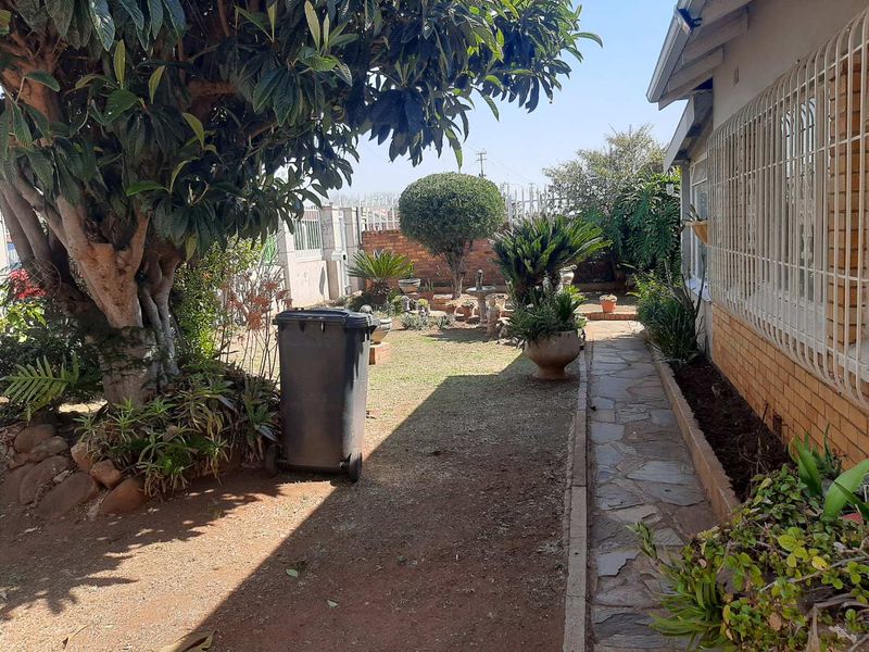 A rear garden area featuring a stone paved walkway, established trees, and a brick boundary wall with security bars on the house windows.