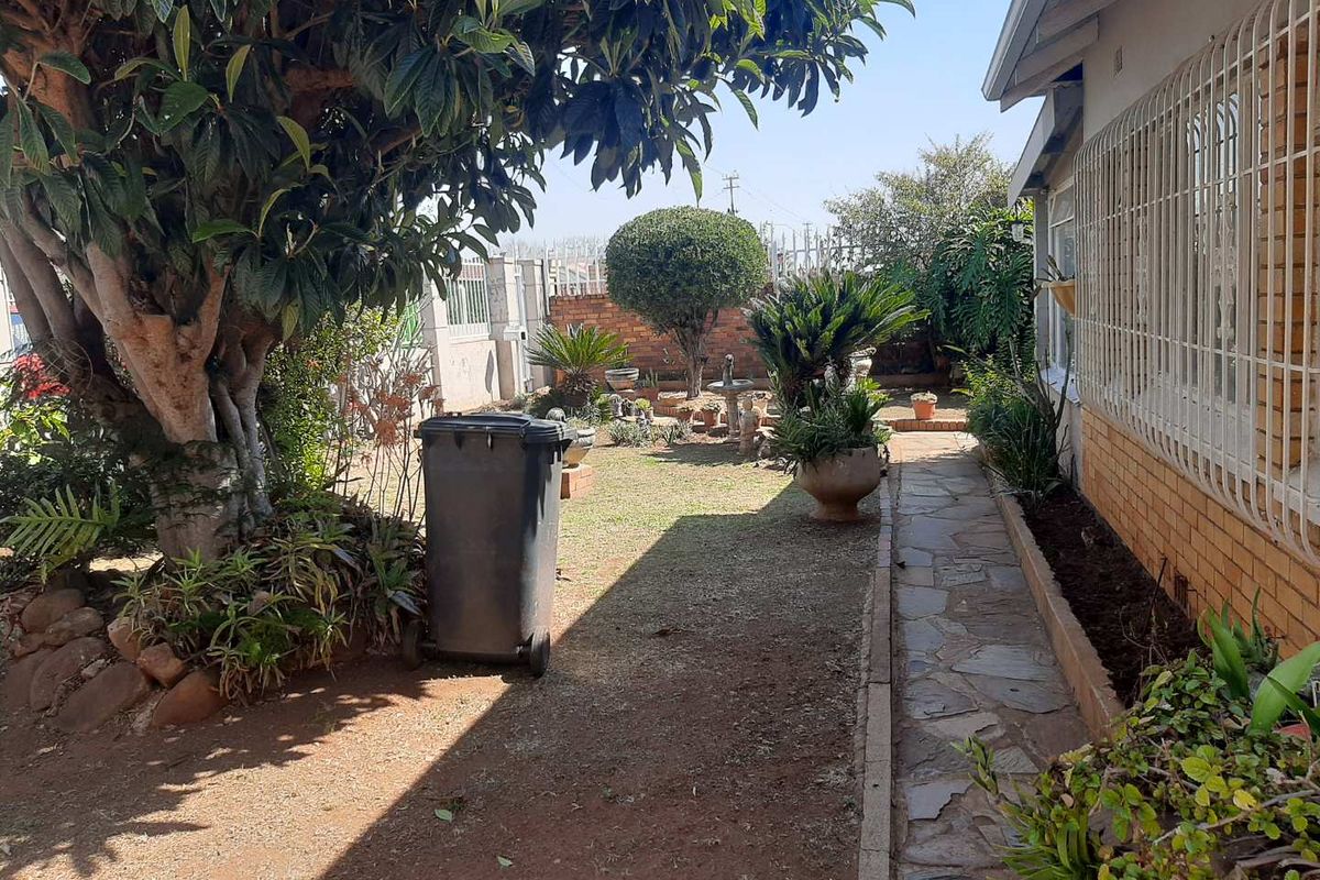 A rear garden area featuring a stone paved walkway, established trees, and a brick boundary wall with security bars on the house windows.