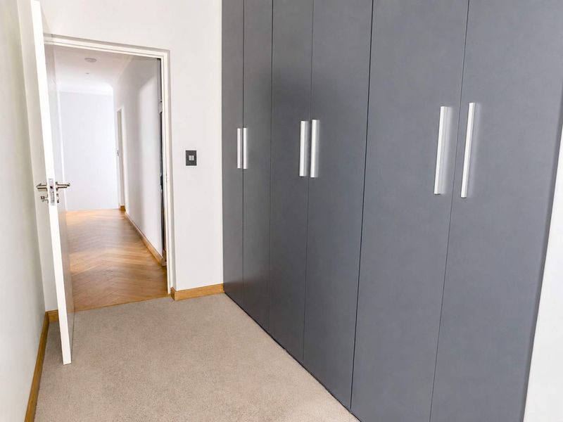 A clean bedroom interior featuring modern grey built-in cupboards, neutral carpeting, and a view into a hallway with herringbone wood flooring.
