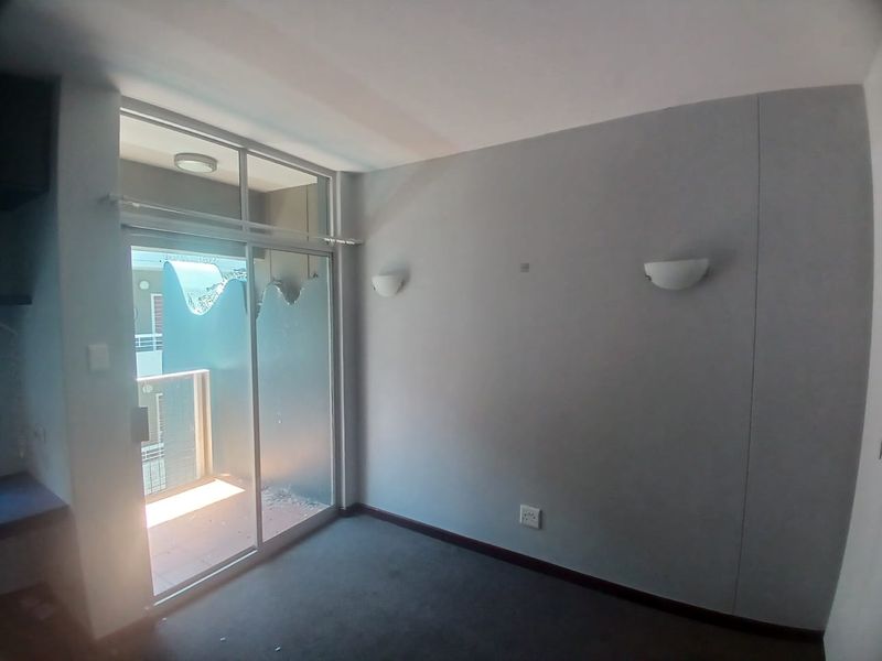 An empty bedroom featuring carpet flooring, wall-mounted lighting, and a sliding glass door that currently has significant damage to the glass panel.
