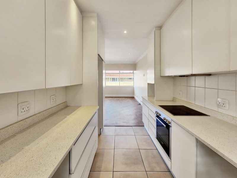 A clean, galley-style kitchen featuring white cabinetry, stone countertops, integrated electric hob and oven, and neutral tiled flooring, leading into an adjacent living area.