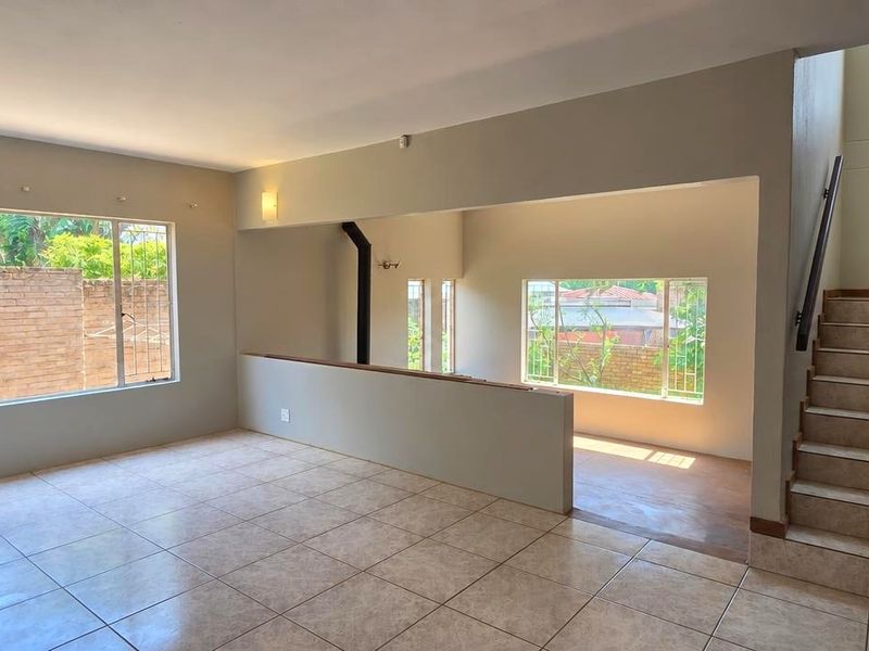 Spacious, open-plan living area featuring tiled flooring, a staircase with a wooden handrail, and large windows with security bars.