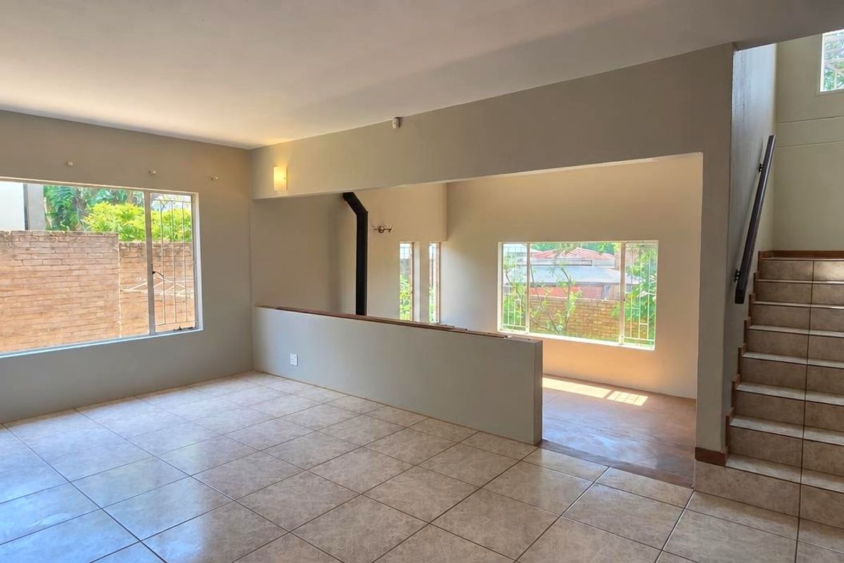 Spacious, open-plan living area featuring tiled flooring, a staircase with a wooden handrail, and large windows with security bars.