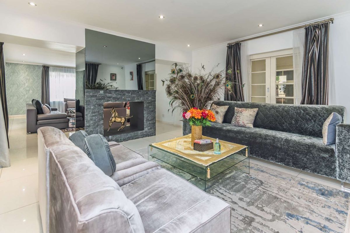 Spacious living room featuring a modern stone-clad fireplace with a mirrored surround, recessed lighting, and tiled flooring, leading to an adjacent seating area.