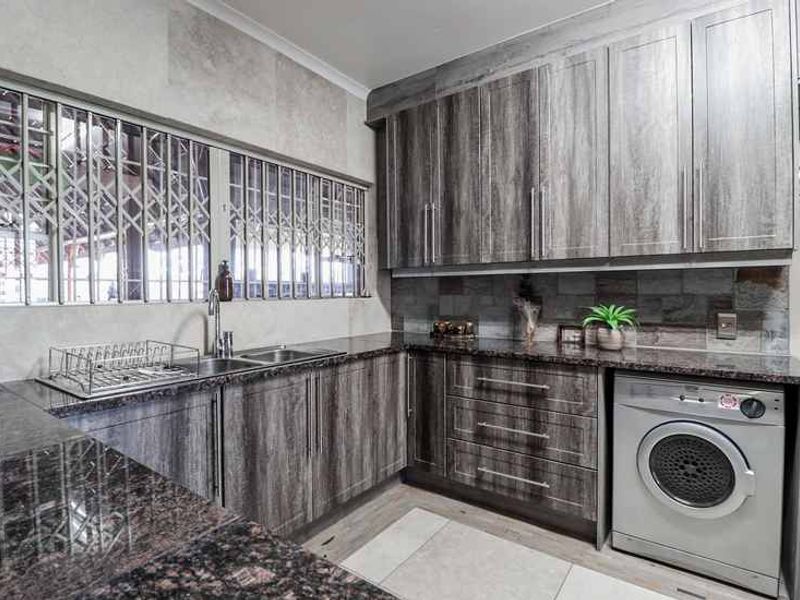 A well-maintained scullery featuring modern wood-grain cabinetry, granite countertops, a stainless steel sink, and a tiled backsplash with integrated laundry appliance space.