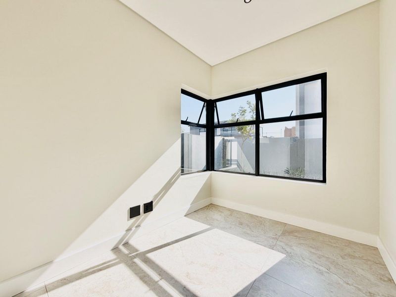 A bright, modern, and empty bedroom featuring large corner windows with black aluminium frames and neutral tiled flooring.