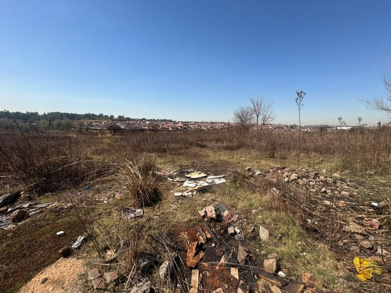 A view of a vacant residential plot featuring dry, overgrown vegetation and scattered debris, with a distant view of the surrounding neighborhood.