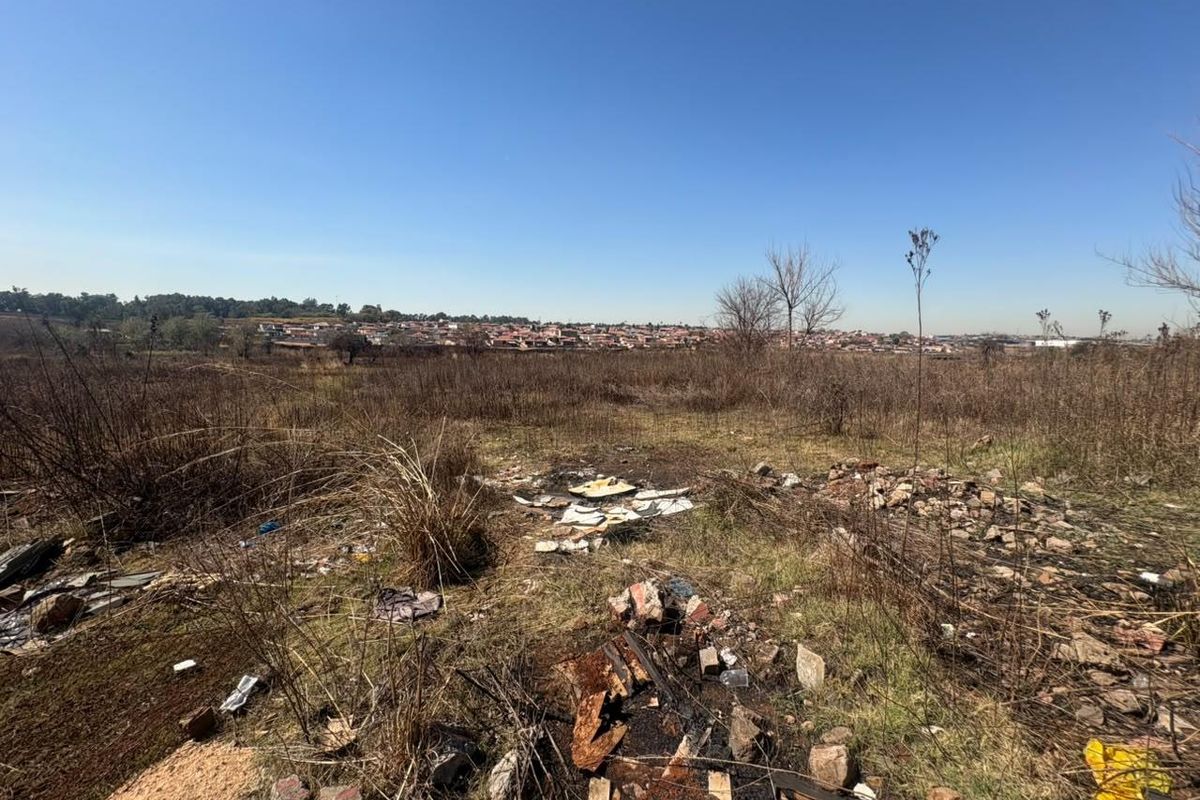 A view of a vacant residential plot featuring dry, overgrown vegetation and scattered debris, with a distant view of the surrounding neighborhood.